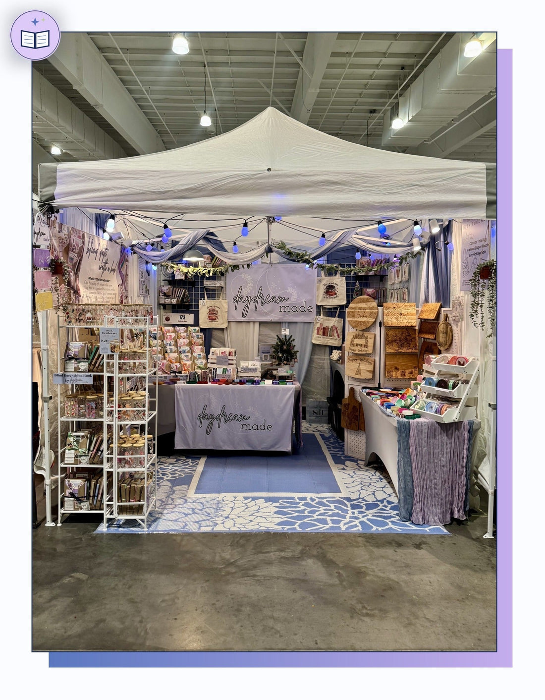 Daydream Made craft fair booth setup inside a 10x10 pop-up tent with folding metal shelving, grid walls, string lights, a custom table cover, and a blue rug