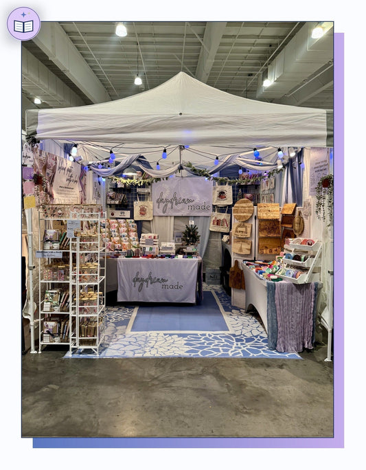 Daydream Made craft fair booth setup inside a 10x10 pop-up tent with folding metal shelving, grid walls, string lights, a custom table cover, and a blue rug