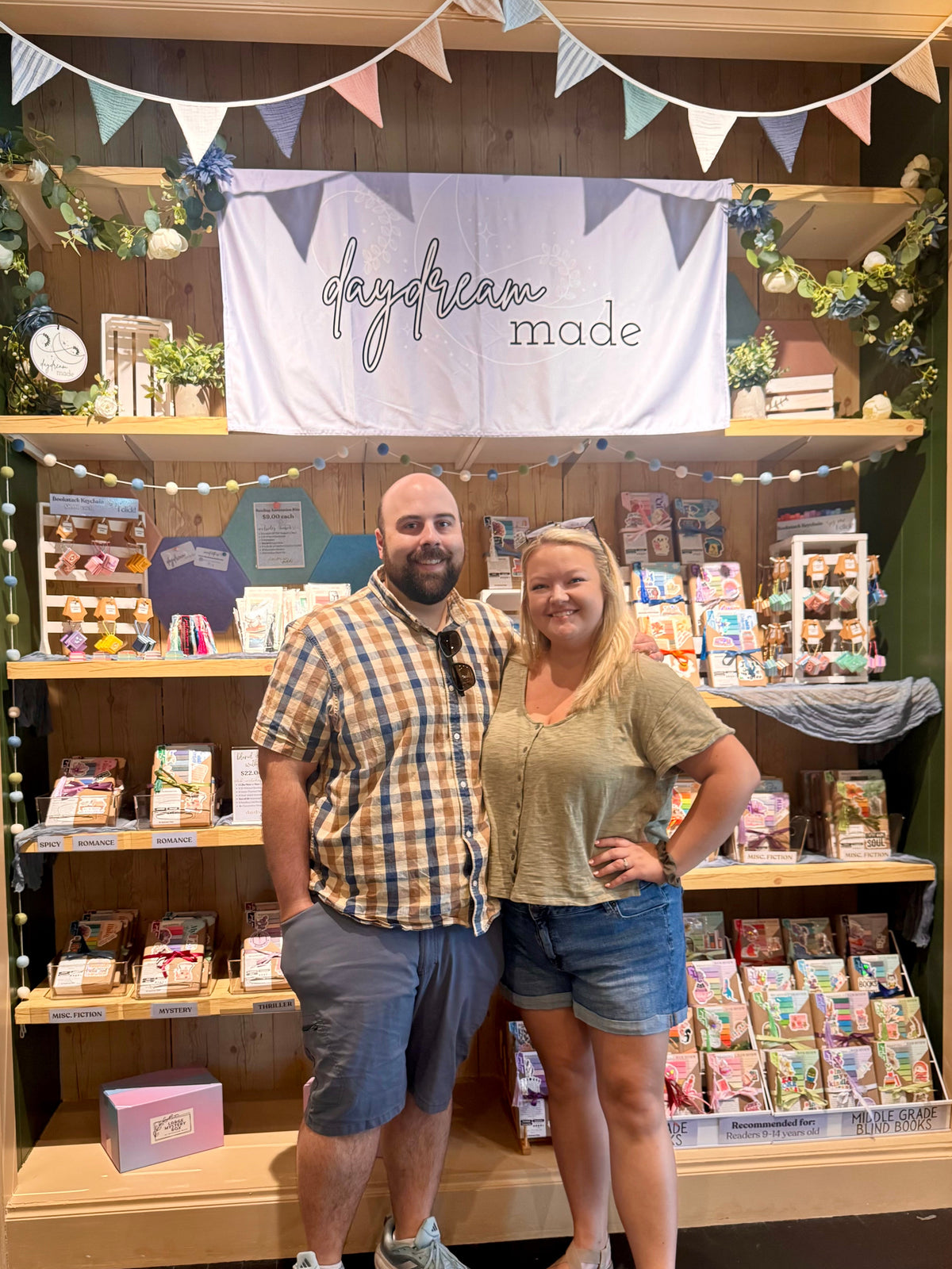 A man and a woman stand smiling in front of a retail display shelf decorated with bunting, greenery, and a white banner reading 'daydream made'. The shelves contain assorted packaged goods and small decor items.